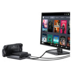 En sort GPD WIN 5 håndholdt konsol vises docket og forbundet via kabler til en skærm. Skærmen viser en Steam OS-lignende grænseflade med et gitter af spiltitler som Dead Cells, Deathloop, Portal 2 og Stardew Valley, hvilket illustrerer enhedens evne til at drive en stationær spiloplevelse. 